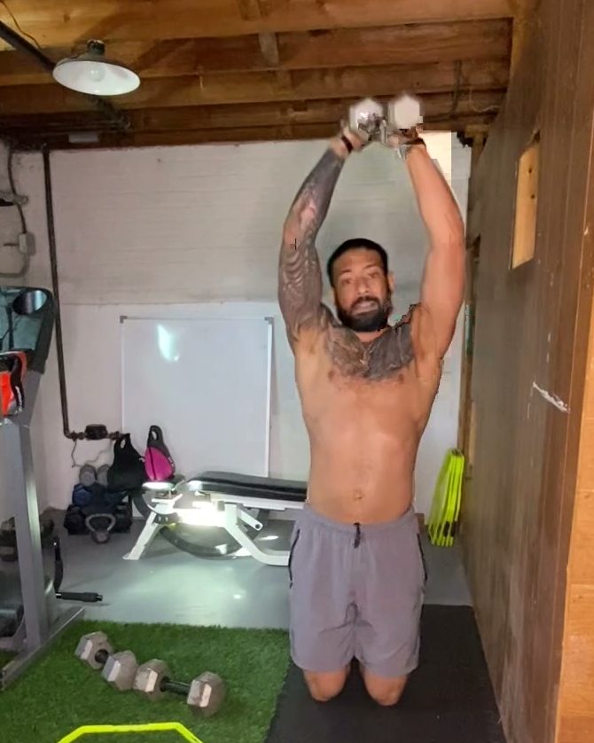 Eddie kneeling and pressing two dumbbells overhead in his home gym